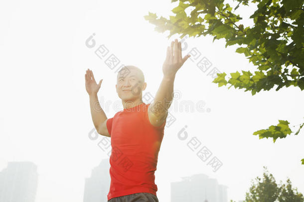 中国北京，一位身穿红衫、笑容满面、肌肉发达的年轻男子在树旁伸展双臂