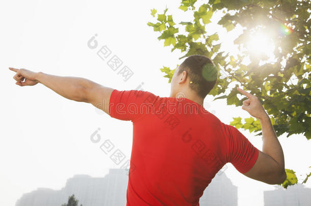 中国北京，年轻肌肉男在树旁伸展双臂手指朝天的后视图