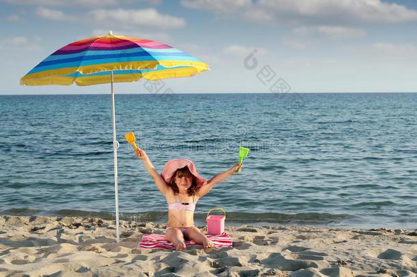 小女孩双手举着坐在沙滩上的遮阳伞下