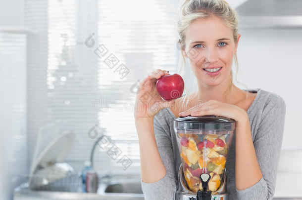 快乐的女人倚在装满水果的榨汁机上，手里拿着红苹果