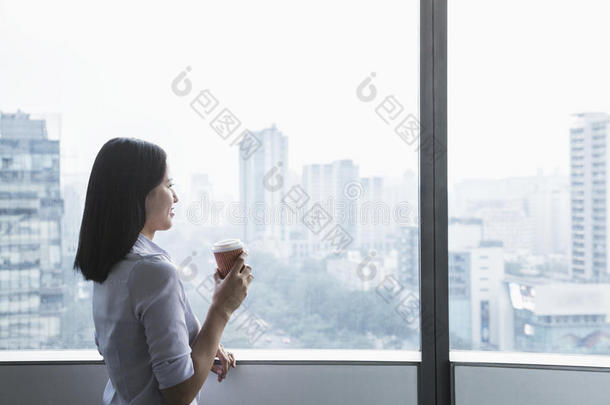 微笑的年轻女商人端着咖啡杯，望着窗外，中国北京的城市风景