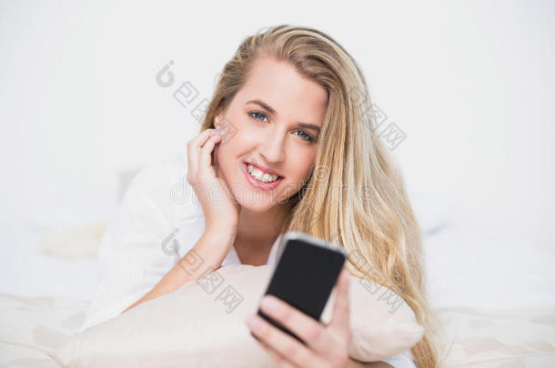 微笑的模特拿着智能手机躺在舒适的床上