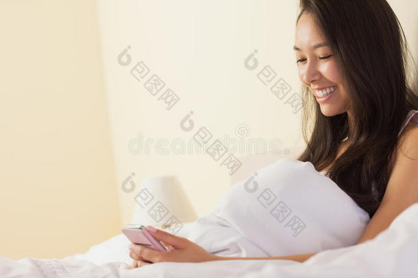 快乐的亚洲年轻女子坐在床上用智能手机