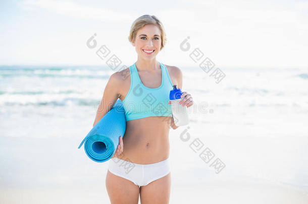 一位<strong>身穿运动服</strong>的金发微笑女士，手里拿着一个瓶子和一张运动垫
