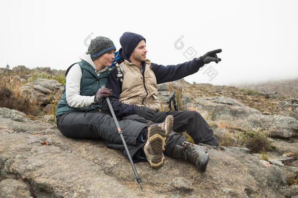徒步旅行时，一对夫妇坐在岩石上，拄着登山杖