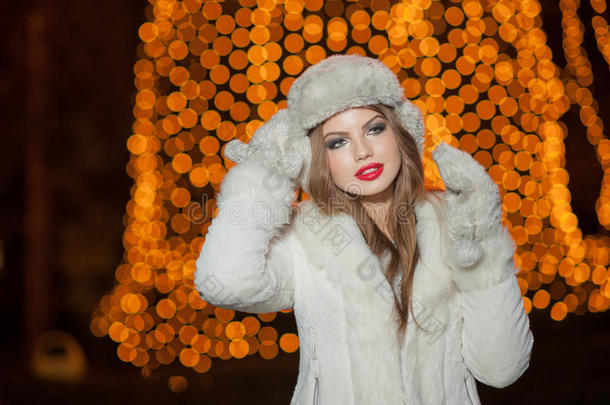 一位时尚的女士，穿着白色毛皮饰品，在户外，背景是明亮的圣诞灯。年轻美女画像