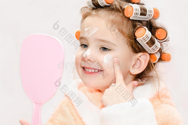 快乐可爱的小女孩，戴着卷发器，涂着镜子用的面霜