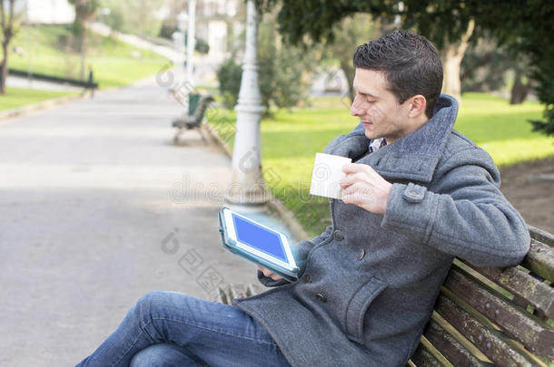 坐在木头长凳上，拿着平板电脑和咖啡杯的男人