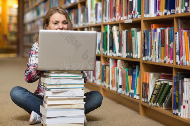快乐的学生坐在图书馆地板上用笔记本电脑放在一堆书上