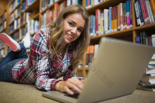 用笔记本电脑躺在图书馆地板上微笑的年轻学生