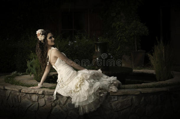 美丽的新娘穿着无肩带复古婚纱斜靠在庭院喷泉旁