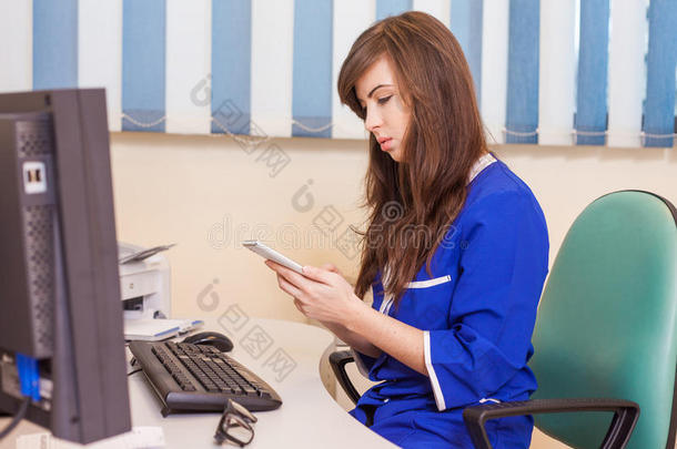 女<strong>医生</strong>在他的电脑上工作，有白色手机。 她穿着蓝色工作服（外科<strong>医生制服</strong>）。
