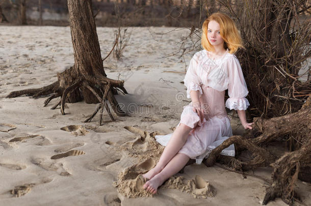 年轻的红头发女孩穿着粉红色的裙子坐在海滩上