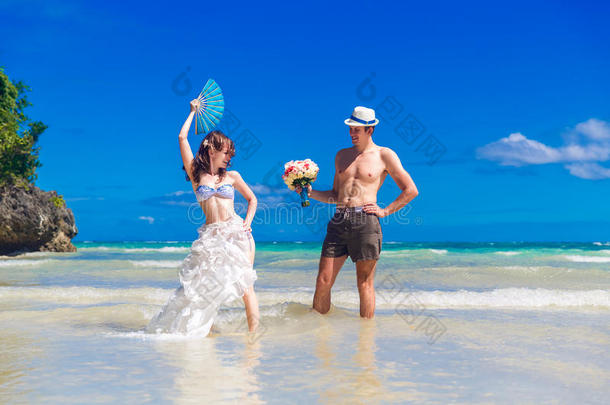 新娘和新郎带着扇子和结婚花束在热带岛屿的海滩上玩得开心。