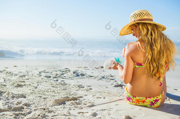 美丽的女人坐在海滩上，戴着遮阳帽，涂着防晒霜