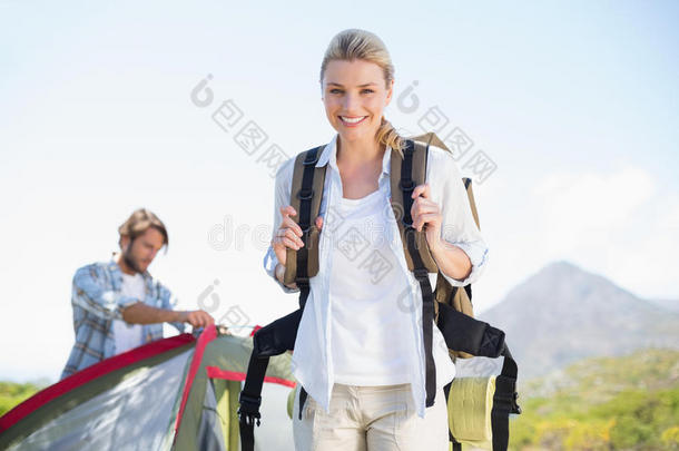 迷人的徒步旅行金发女郎在镜头前微笑，而合作伙伴则在帐篷里