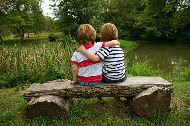 可爱的<strong>双胞胎</strong>小<strong>兄弟</strong>坐在一张木凳上，拥抱在一起，看着美丽的湖水