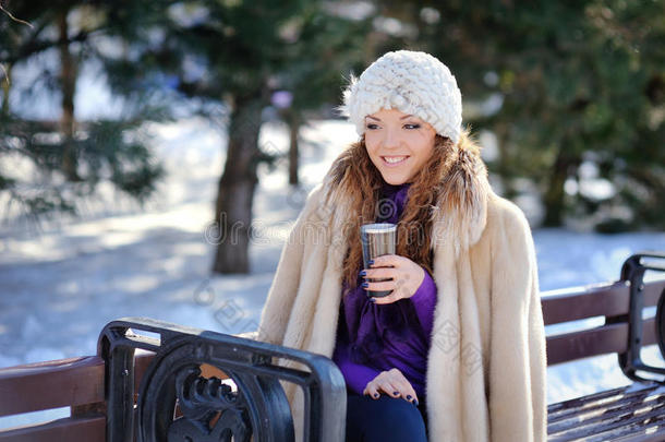 冬天，黑发女孩戴着白帽子，拿着一个杯子坐在长椅上