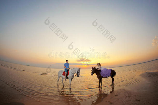 两个骑马的人在海滩上日落时骑马。情侣骑马。年轻漂亮的男人和女人在海上骑马。