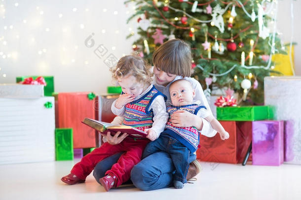 可爱的快乐男孩在一个有圣诞树的黑暗房间里给他蹒跚学步的妹妹和刚出生的弟弟<strong>朗读</strong>