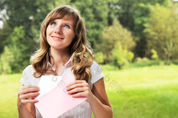 站在模糊的绿色夏日背景上的年轻白人妇女。手里拿着一封粉红色信封里的空白信。信和信封公司
