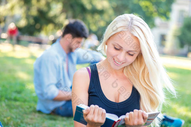 准备测试。当她的朋友坐在草地上时，漂亮的女学生读课本，对着镜头微笑