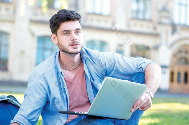 困难的大学。 可爱的男学生拿着笔记本电脑读着一本书，坐在附近的草地上<strong>看</strong>着<strong>远方</strong>
