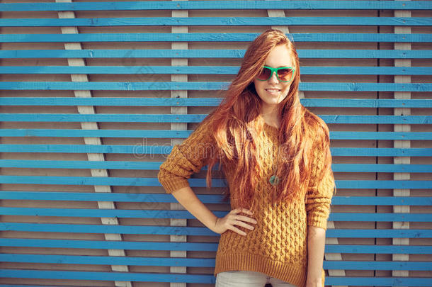 美丽的年轻红发少女戴着墨镜站在蓝色木板墙上夏日温暖