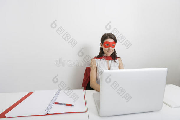 年轻的女商人打扮成超级英雄在<strong>办公</strong>室<strong>办公</strong>桌旁<strong>用笔</strong>记本电脑
