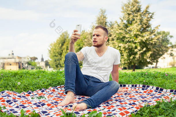 大胡子男人坐在公园里的毯子上。他在用手机