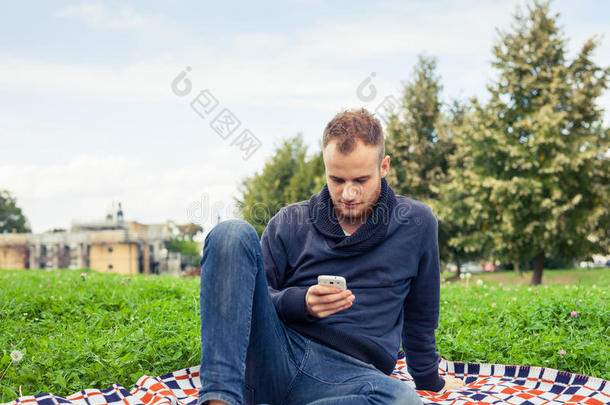 大胡子男人坐在公园里的毯子上。他在用手机