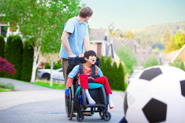 高加索人的父亲帮助残疾的混血儿坐在轮椅上在街上户外踢足球