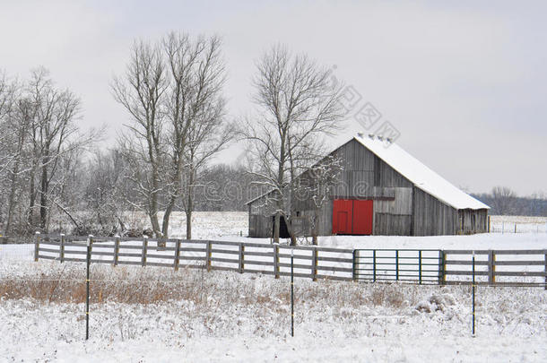 灰色的乡村谷仓，在白雪覆盖的景观上有一扇红色的门