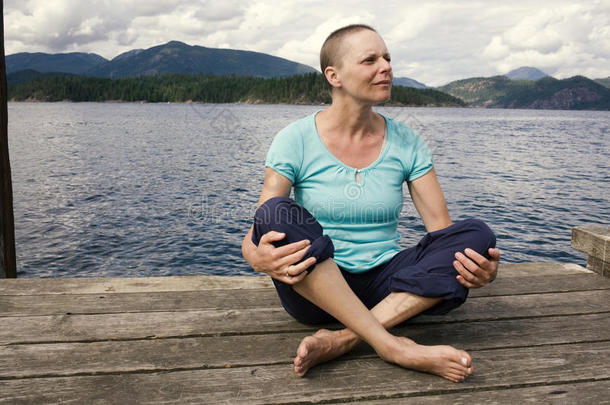 一因化疗而脱发的女人坐在外面的码头上，身后是海洋和山脉