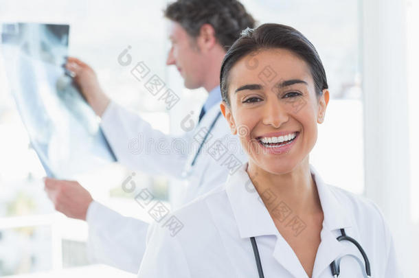 女医生在镜头前微笑，而她的同事在看X光