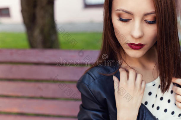 漂亮时尚的女孩穿着黑色皮夹克，黑色口红和化妆在城市的长凳上