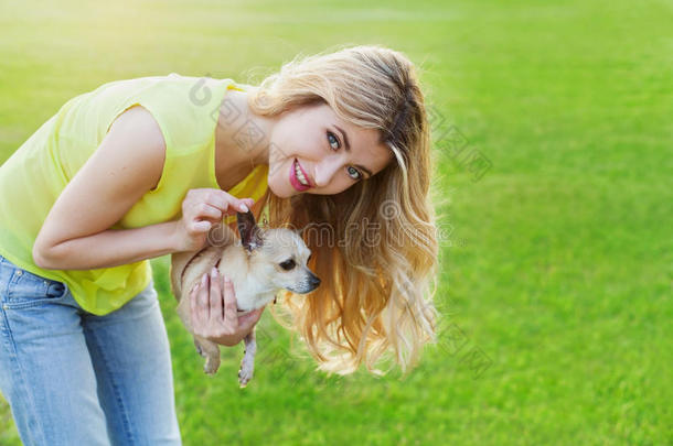 迷人的快乐微笑的女孩或女人抱着可爱的吉娃娃小狗在绿色的草坪上日落