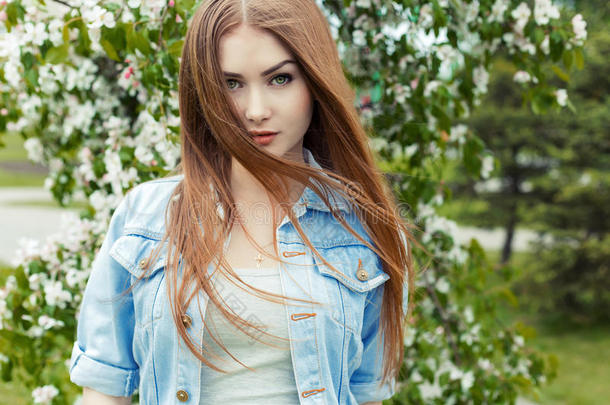 美丽可爱的甜美女孩，留着长长的红色头发和绿色的眼睛，穿着牛仔夹克，靠近公园里的一棵开花树