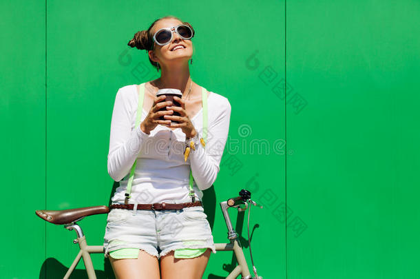 时尚美丽的年轻漂亮女孩穿着短裤和T恤和凉爽的太阳镜站在自行车固定装置旁边的绿色墙壁