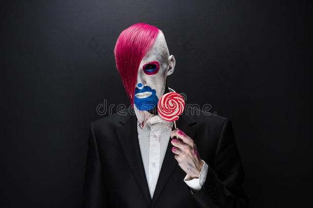 小丑和万圣节主题：可怕的小丑，粉红色的头发，穿着黑色夹克，手里拿着糖果，工作室的黑暗背景上