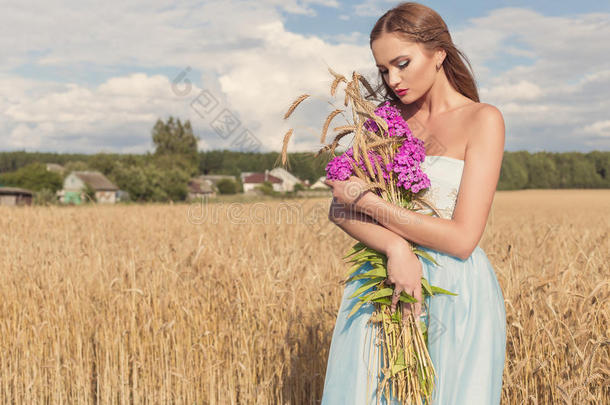 美丽的<strong>苗</strong>条女孩穿着一件蓝色的连衣裙在田野里，在阳光明媚的日落时，他手里拿着一束花和一双<strong>玉米</strong>耳朵