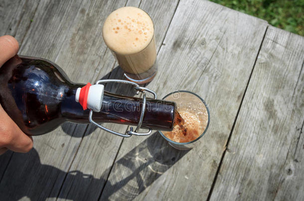泡沫的深色啤酒从夏天花园里的棕色玻璃瓶中倒入高大的玻璃杯中，放在乡村的木桌上