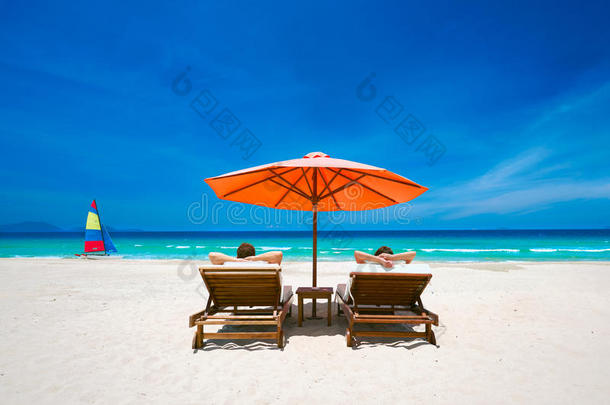 一对夫妇在热带海滩上，在红色雨<strong>伞下</strong>的甲板椅子上。
