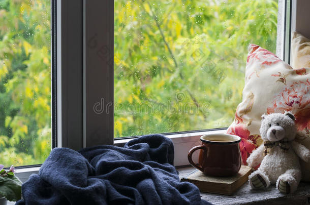 咖啡杯，书，泰迪熊，枕头和一个格子在轻木表面与窗户雨天的看法。 复古风格。