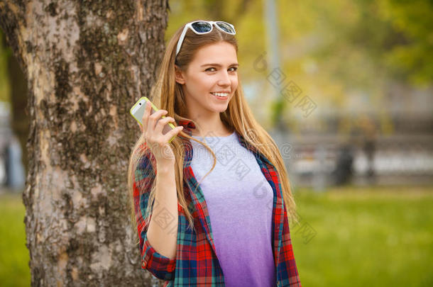 迷人的微笑女孩在夏天的城市公园拿着手机。 现代快乐的女人带着智能手机，户外