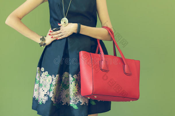 时尚漂亮的大红色手提包在女孩的手臂上，穿着一件时尚的黑色连衣裙，在温暖的夏天在墙上摆姿势