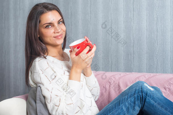 一个女人手里拿着一个红色的杯子，微笑着坐在沙发上，沙发上看着相机