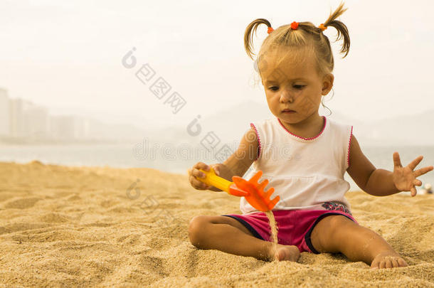 美丽的婴儿坐在面对相机和玩玩具耙在沙滩上的沙子。