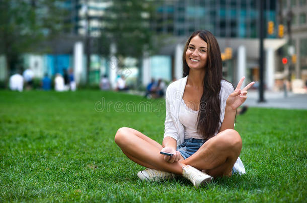 高加索游客年轻的美女坐在城市公园的绿草地上，展示胜利的标志