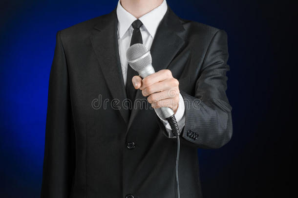 商业与演讲主题：<strong>演播室</strong>里，一个身穿黑色西装的男人在灰深蓝色的隔离背景上拿着麦克风